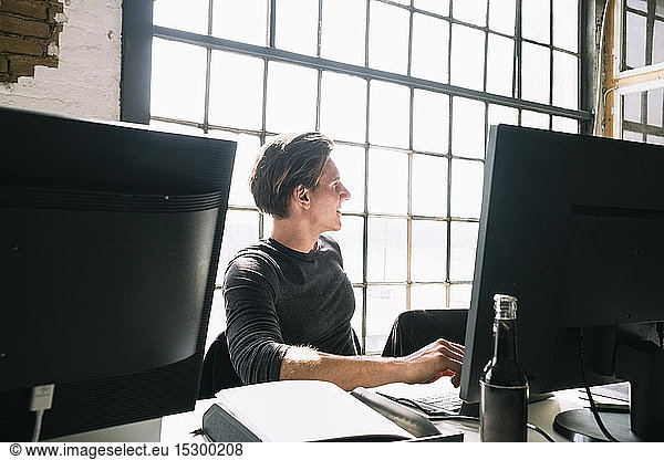 Young male hacker looking away while sitting at computer desk in creative workplace