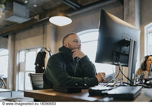 Young male hacker with hand on chin using computer at desk in creative office