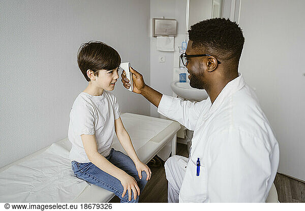 Young male pediatrician measuring temperature of boy using infrared thermometer at clinic
