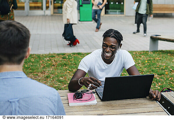 Young male student laughing while sitting with friend in campus