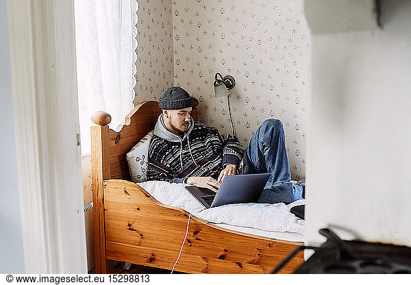Young male using laptop while relaxing on bed seen through doorway at home