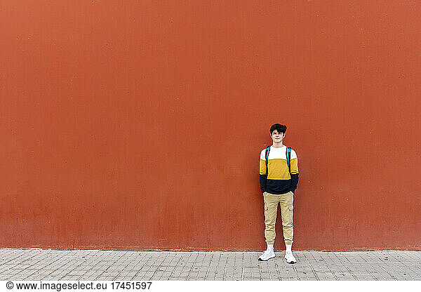 Young male with backpack standing on red wall while looking to camera