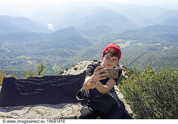 Young male with wool red hat sitting against mountain taking a selfie