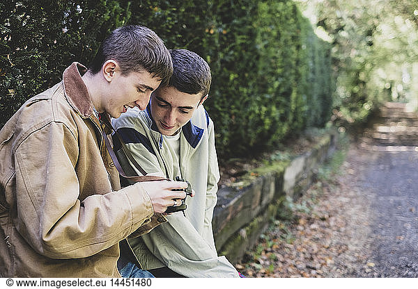 Young man and teenage boy with short brown hair wearing casual jackets sitting on hedged stone wall  looking at image on digital camera.