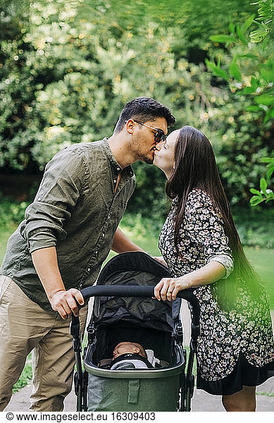 Young man and woman kissing while standing with baby carriage at park