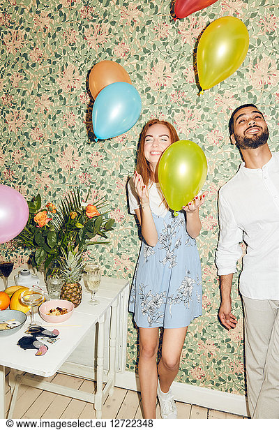 Young man and woman playing with balloons while standing against wallpaper at home during dinner party