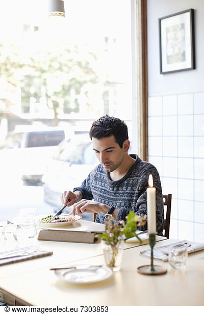 Young man at restaurant table eating meal while using digital tablet