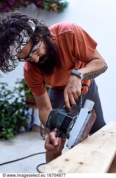 Young man beard polishing a wooden plank with a power sander