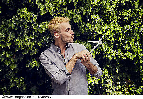 Young man blowing on wind turbine model in front of hedge