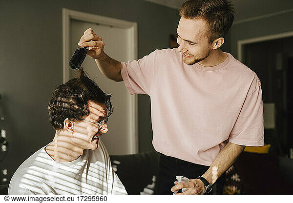 Young man combing male friend's hair while sitting at home