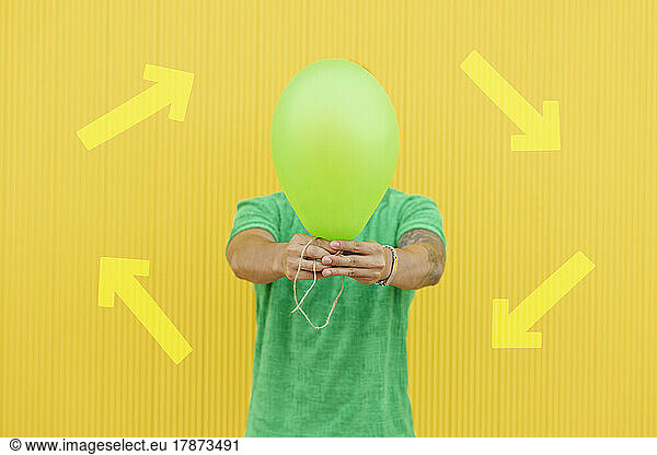 Young man covering face with green balloon in front of arrow symbols on yellow wall