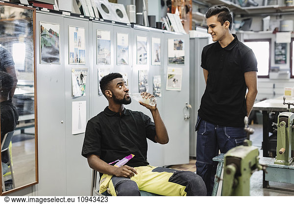 Young man drinking water while looking at coworker in workshop