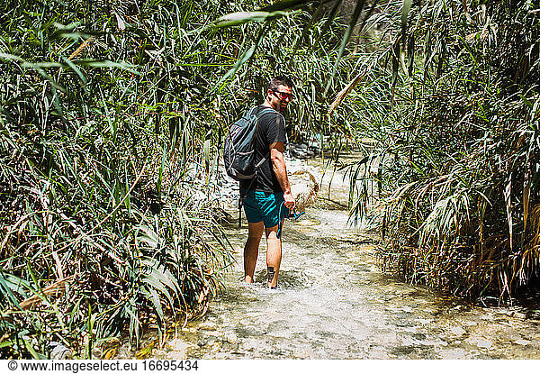 Young man hiking up river surrounded by green plants in summer