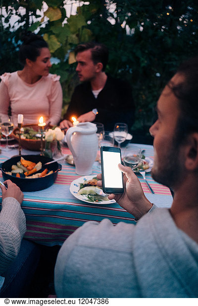 Young man holding mobile phone while friends communicating at dining table