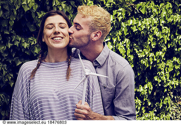 Young man holding wind turbine model kissing girlfriend in front of hedge