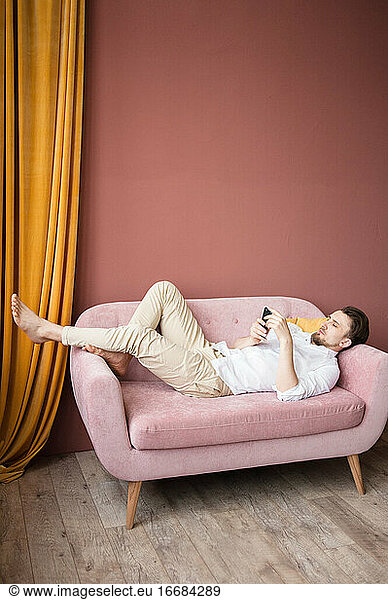Young man in a white shirt works on his phone while lying on pink sofa