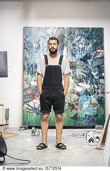 Young man in dungarees standing in artist's sudio  with hands in pockets