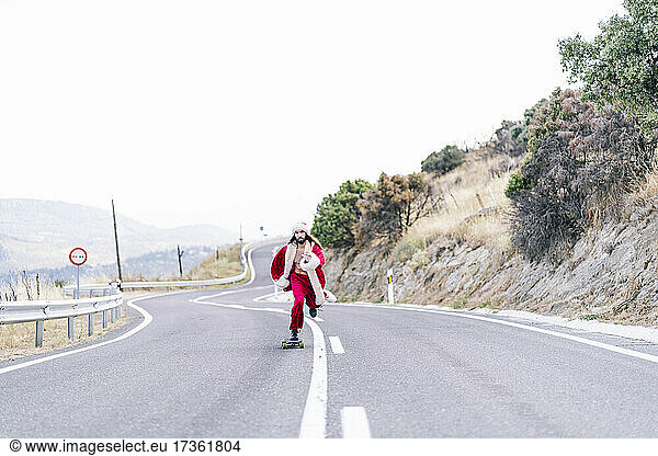 Young man in Santa Claus costume skateboarding on road