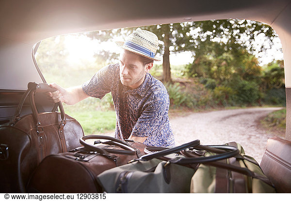 Young man in trilby removing luggage from car boot on rural road