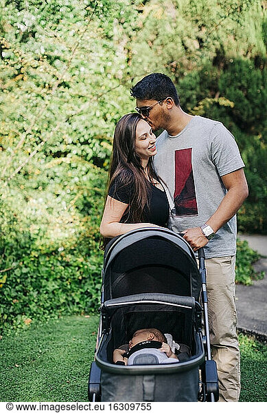 Young man kissing woman while standing with baby carriage at park