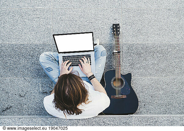 young man long hair writing on laptop sitting next acoustic guitar
