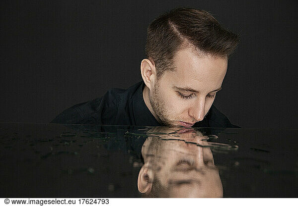 Young man looking at reflection with water on mirror against black background