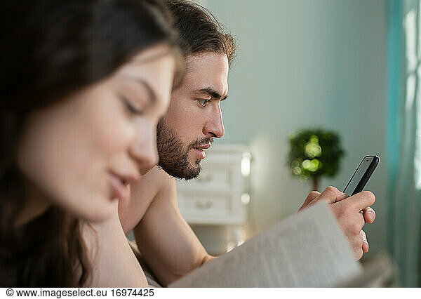 Young Man Looking At Smartphone while his girlfriend Reading Boo