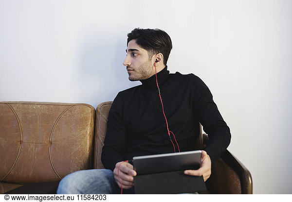 Young man looking away while listening music through digital tablet on sofa at office