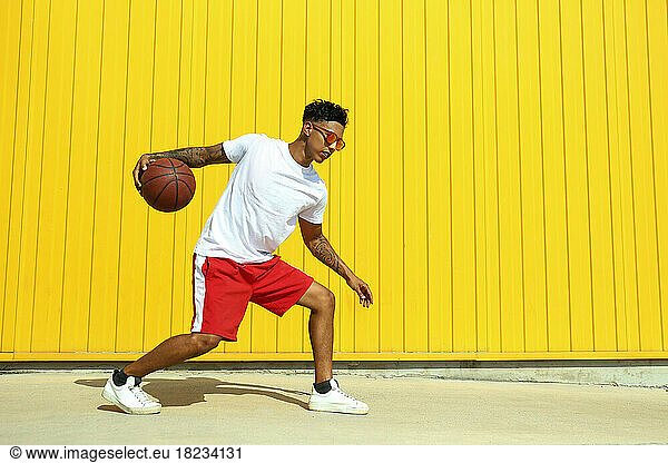 Young man playing with basketball on footpath in front of yellow wall