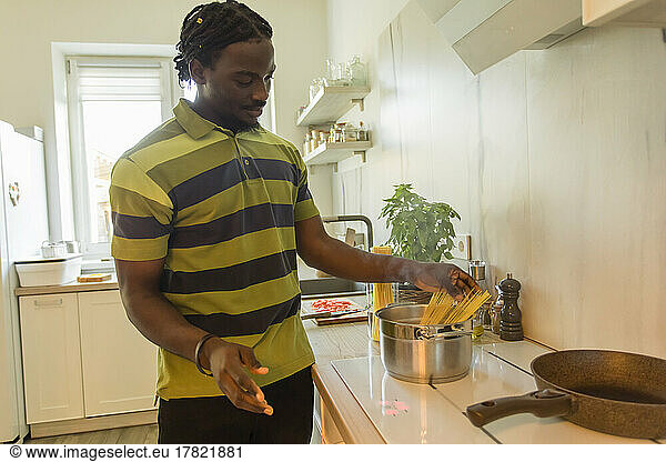 Young man putting uncooked spaghetti in cooking pot at kitchen