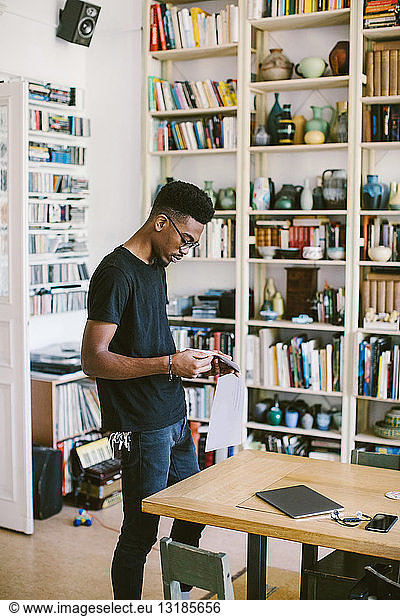 Young man reading paper while standing by table at home