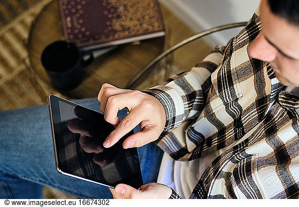 Young man seated at home using digital tablet. Chilling next to the window consuming digital content. Top view.