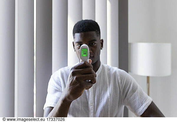 Young man showing temperature on infrared thermometer at home