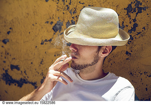 Young man sitting in front of flaking yellow wall  smoking a cigarette