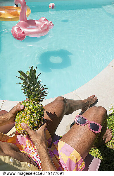 Young man sitting with pineapple and sunglasses near poolside