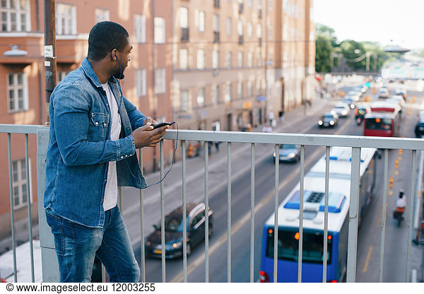 Young man standing with mobile phone on footbridge while looking at city street