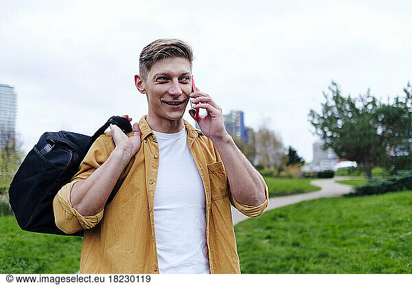 Young man talking on smart phone carrying bag at park