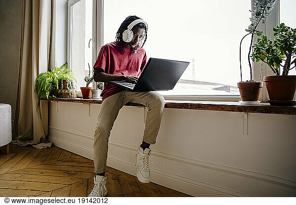 Young man using laptop sitting on window sill at home