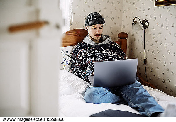 Young man using laptop while relaxing on bed seen through doorway at home