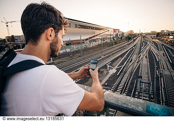 Young man using mobile phone while leaning on railing over railroad tracks in city