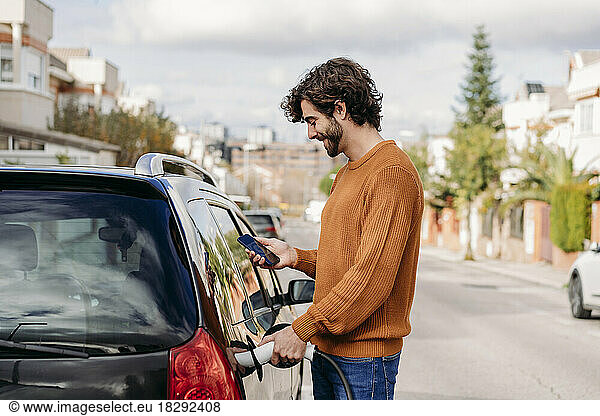 Young man using smart phone and charging car at electric vehicle charging station