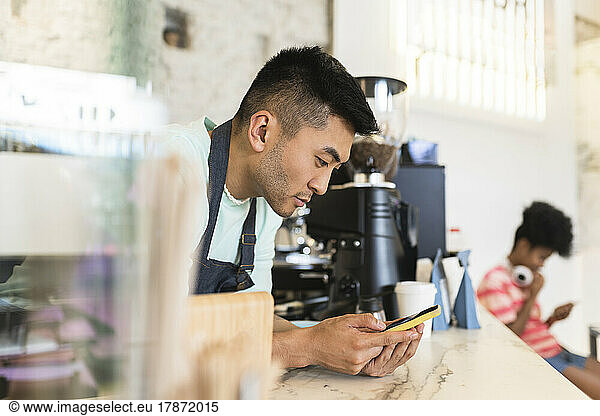 Young man using smart phone leaning on checkout in coffee shop