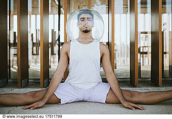 Young man wearing glass container on head with legs apart meditating against built structure
