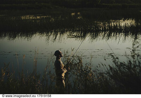Young man wearing hat fly fishes on shore of creek in setting sun