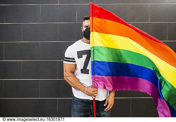 Young man wearing mask holding rainbow flag while standing against wall