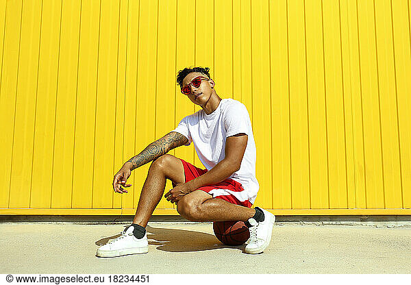 Young man wearing red sunglasses sitting on basketball in front of yellow wall