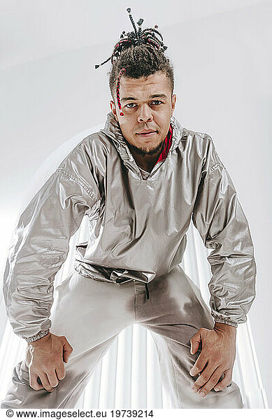Young man wearing silver colored clothing bending with hands on knee in front of white wall