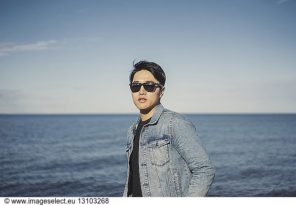 Young man wearing sunglasses and denim jacket while standing against sea and sky
