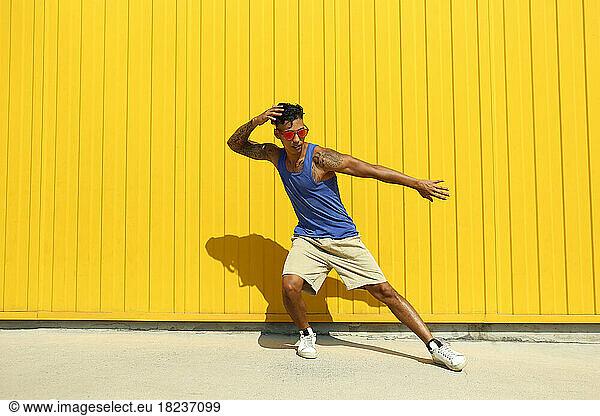 Young man wearing sunglasses dancing on footpath in front of yellow wall