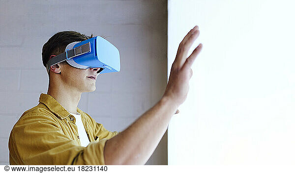 Young man wearing virtual reality headset gesturing by white wall
